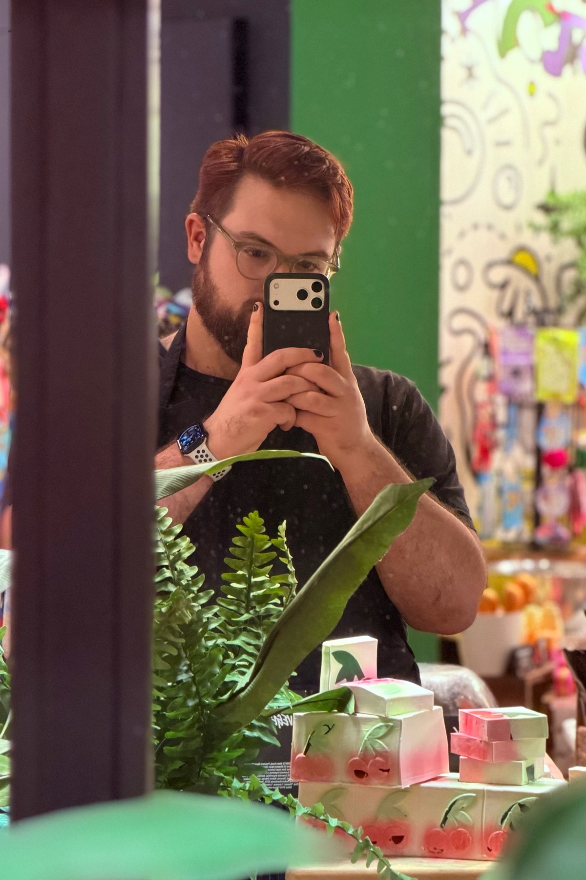 A man with red hair, a beard, and glasses takes a mirror selfie while holding his phone. He is wearing a black t-shirt and is surrounded by plants and cherry-themed items.
