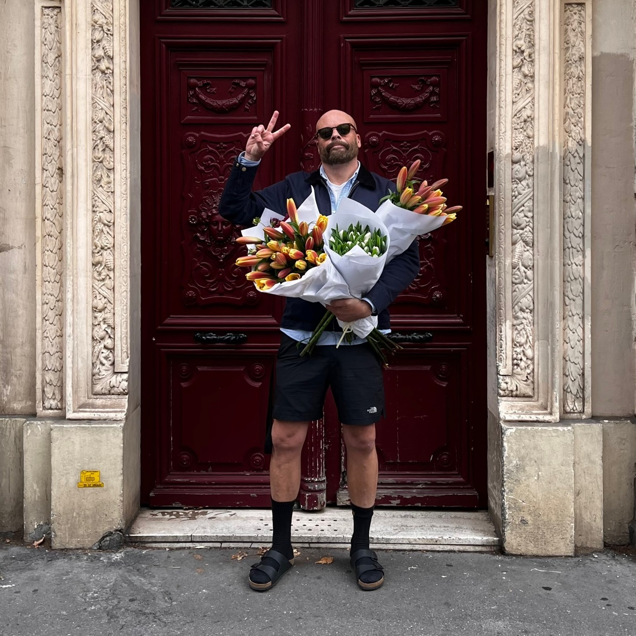 A bald man with a beard and sunglasses stands in front of a large red door, holding two bouquets of tulips and making a peace sign with his hand.