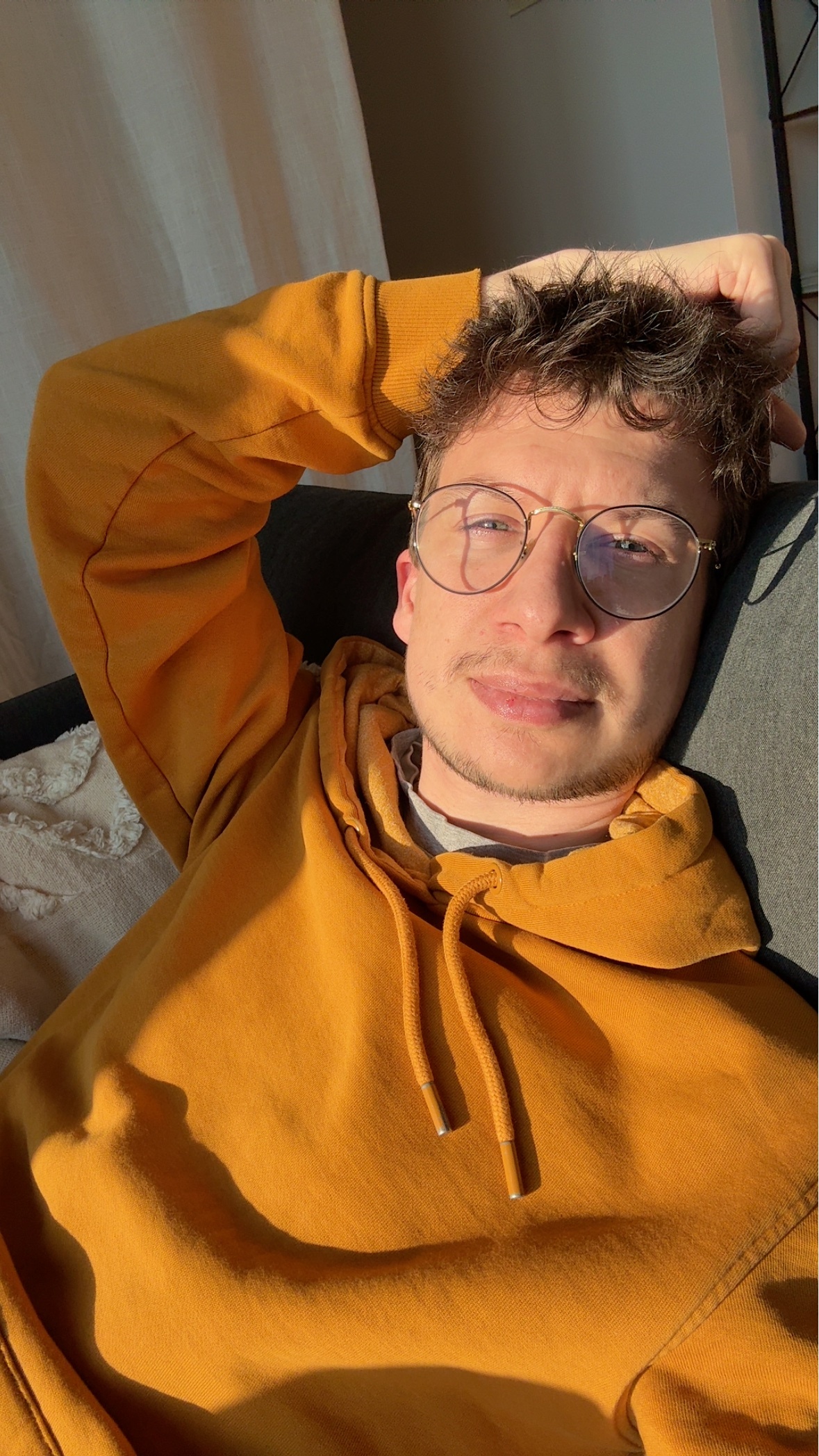 A man wearing glasses and a yellow hoodie poses for a selfie in the sunlight. The photo is taken from a low angle.