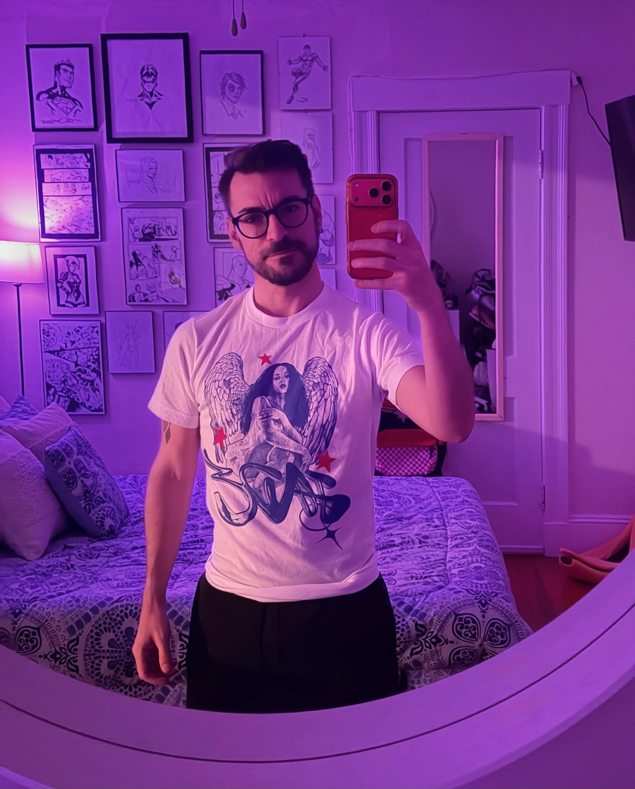 A man takes a selfie in a bedroom with a mirror. He is wearing a graphic t-shirt and glasses.