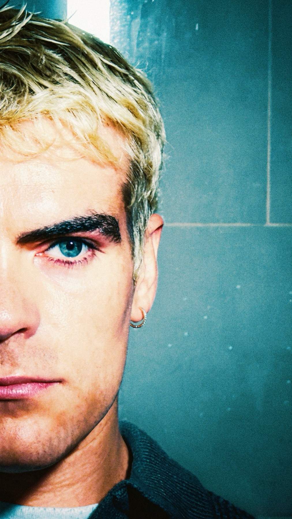 A close-up shot of a man's face with blonde hair and striking blue eyes. He is wearing subtle makeup and a small hoop earring.