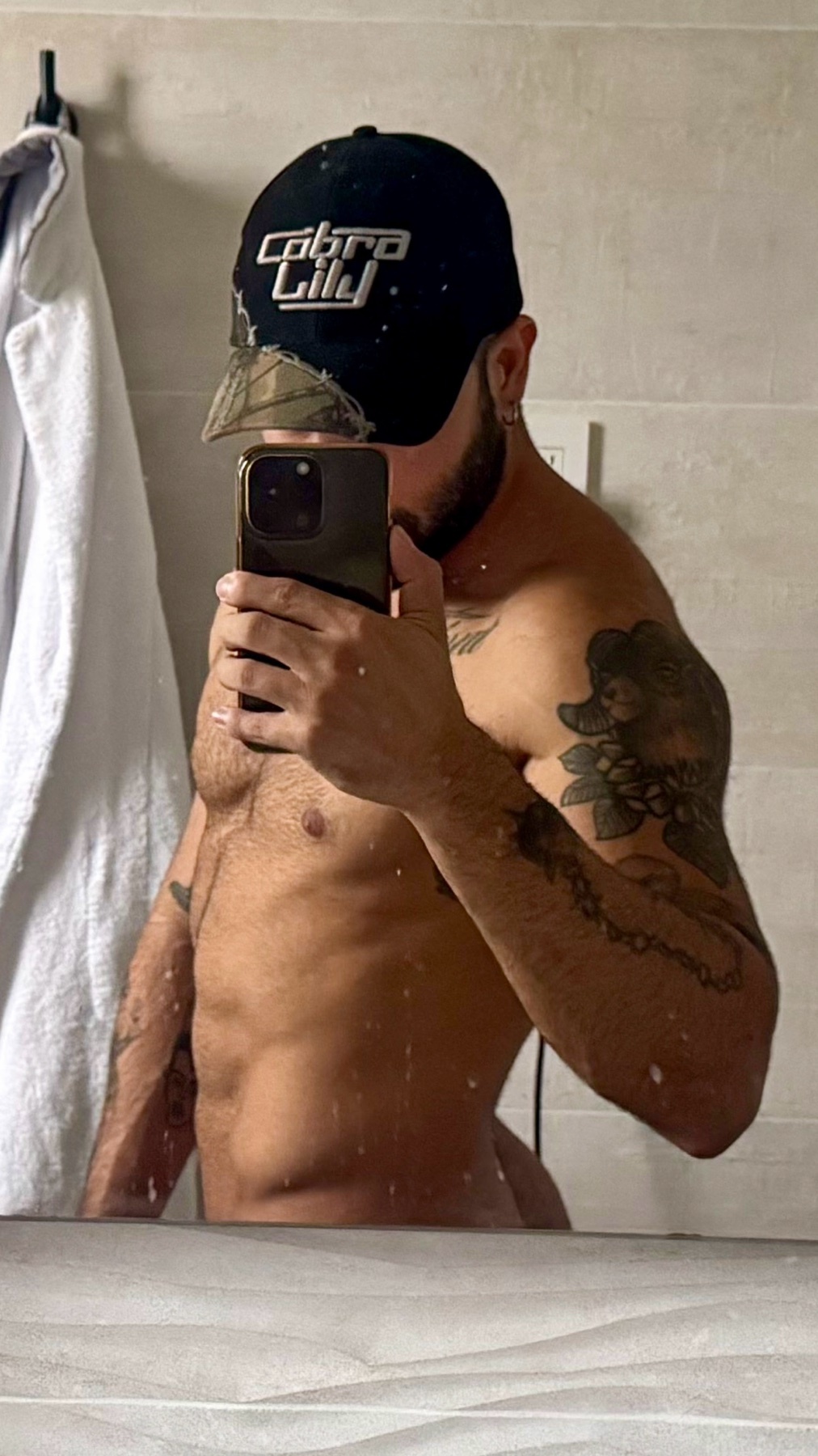 A man takes a mirror selfie in a bathroom, showcasing his physique. He is shirtless, wearing a cap, and has tattoos.