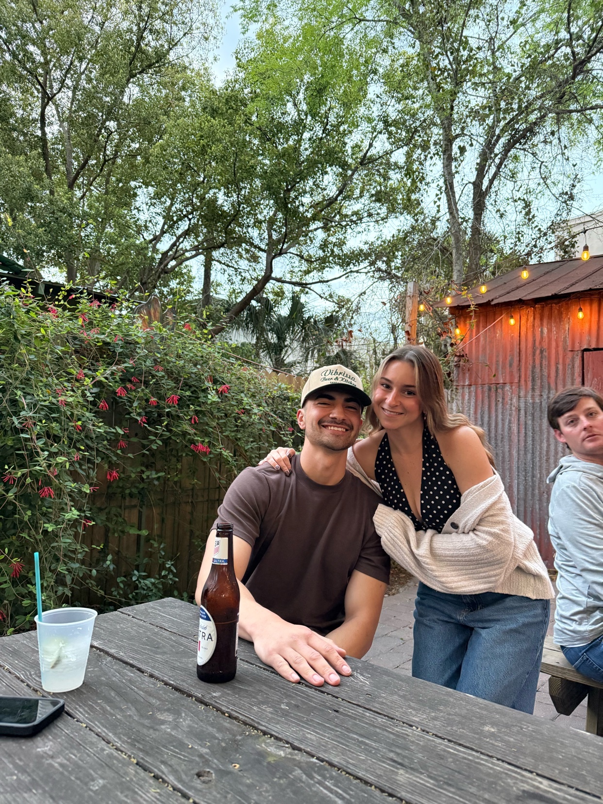 A man and woman smile at the camera while sitting at a picnic table outdoors. The man wears a brown t-shirt and a baseball cap, while the woman wears a black and white polka dot top with a cream cardigan.