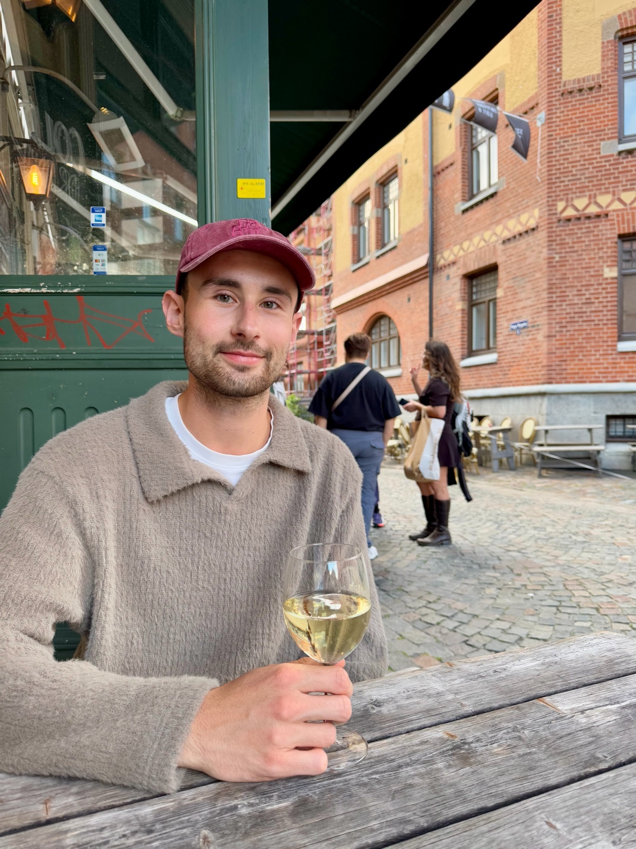 A man sits at an outdoor restaurant table, holding a glass of wine. He is smiling at the camera, with a cityscape in the background.
