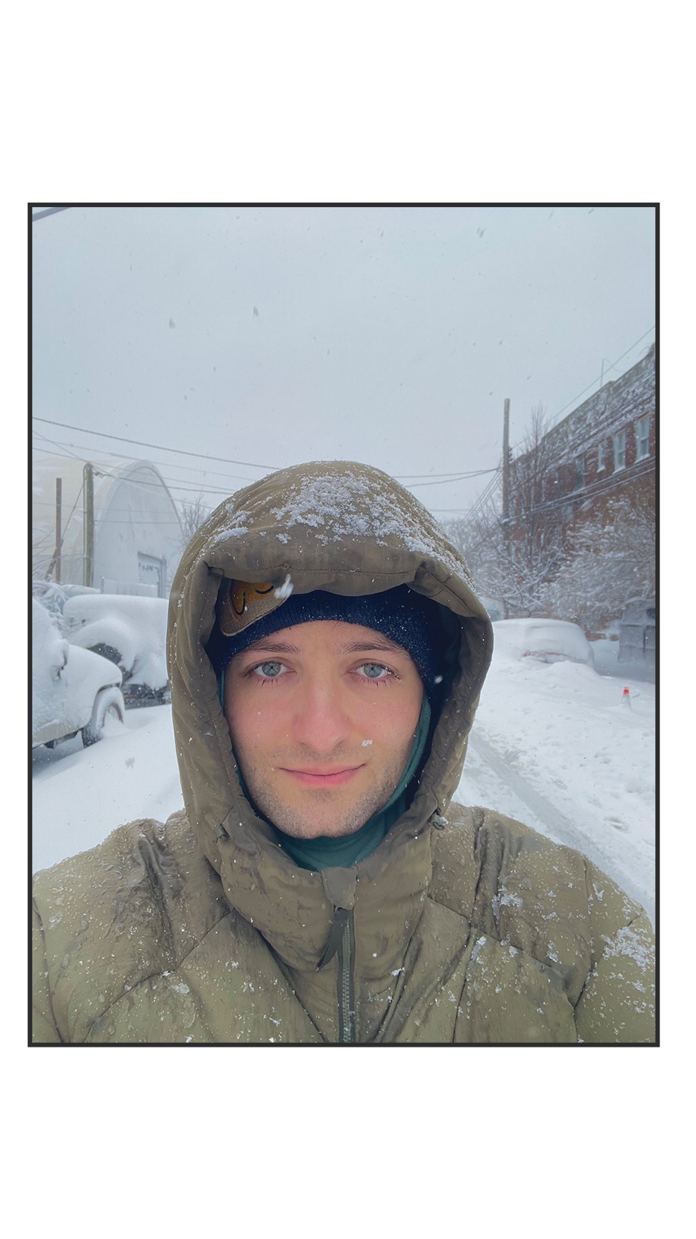 A man takes a selfie in the snow, bundled up in a hooded jacket. The background shows a snowy street and parked cars.
