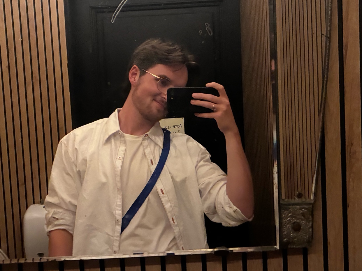 A man takes a mirror selfie in a bathroom. He is wearing a white button-down shirt over a white t-shirt and has a blue strap across his chest.
