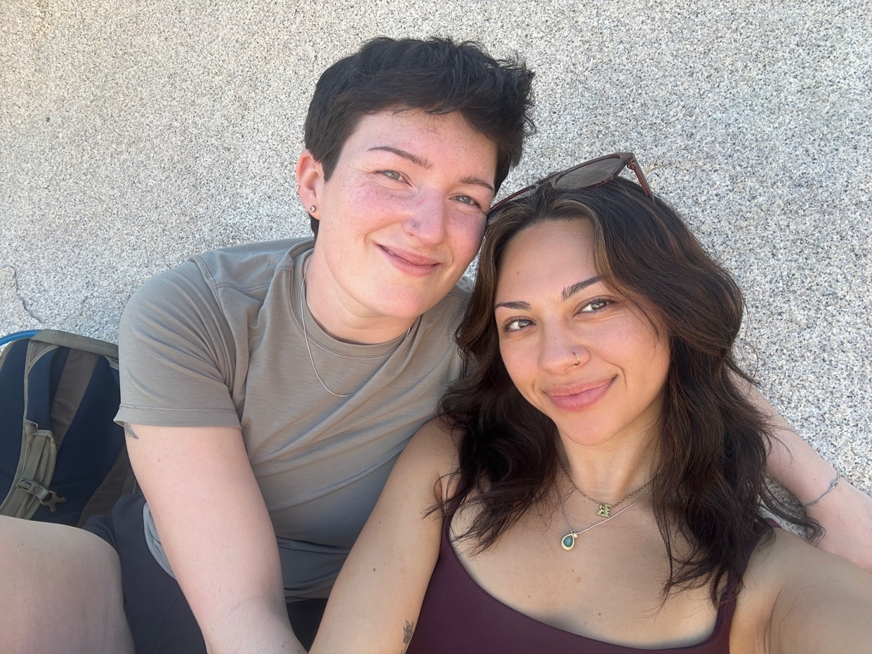 A non-binary person and a woman smile for a selfie outdoors. The non-binary person has short, dark hair and freckles, while the woman has long, dark hair and dimples.