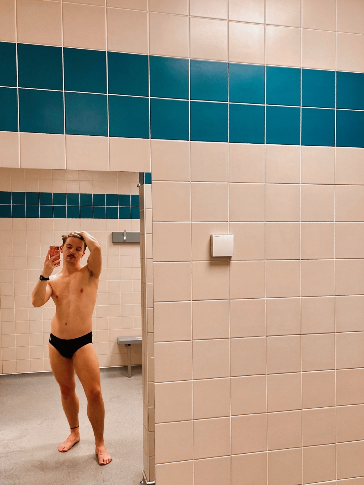 A man takes a mirror selfie in a gym locker room. He is shirtless and wearing black underwear.