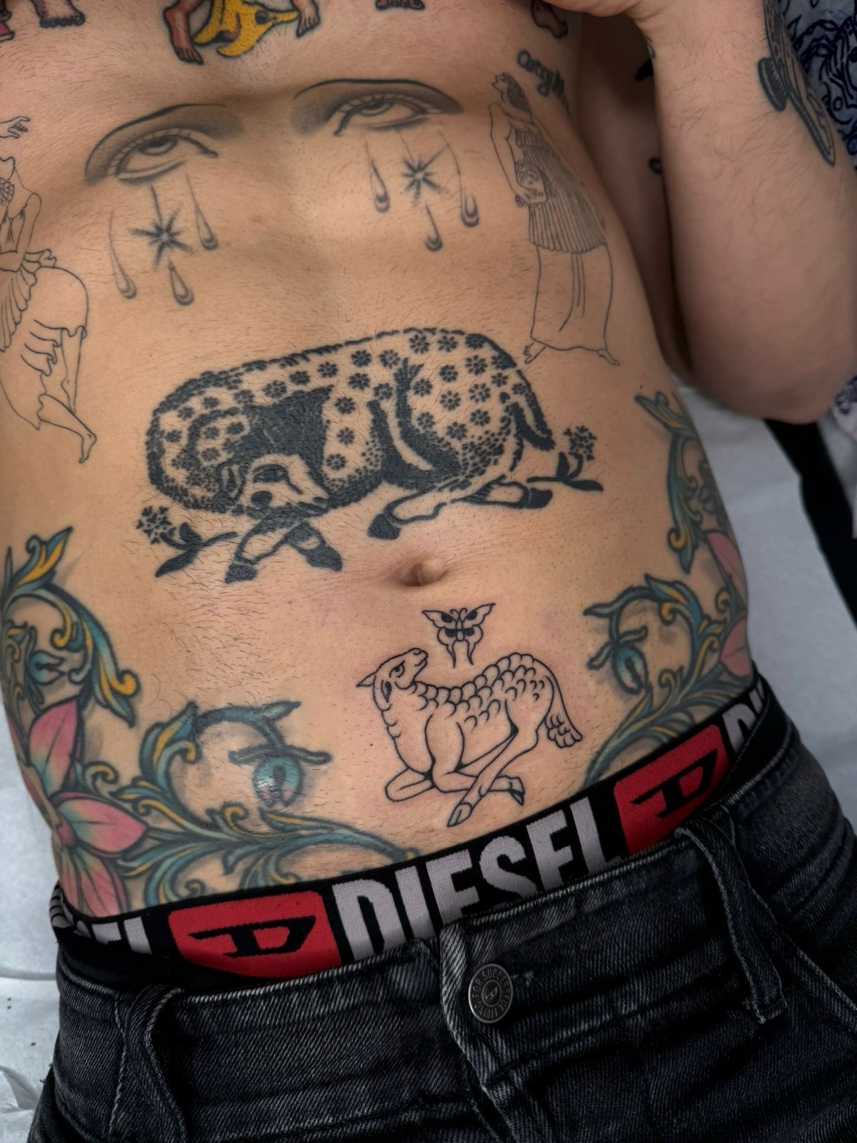 A close-up shot of a person's torso covered in various tattoos, including a leopard, a lamb, and floral designs. The person is wearing black jeans and Diesel underwear.