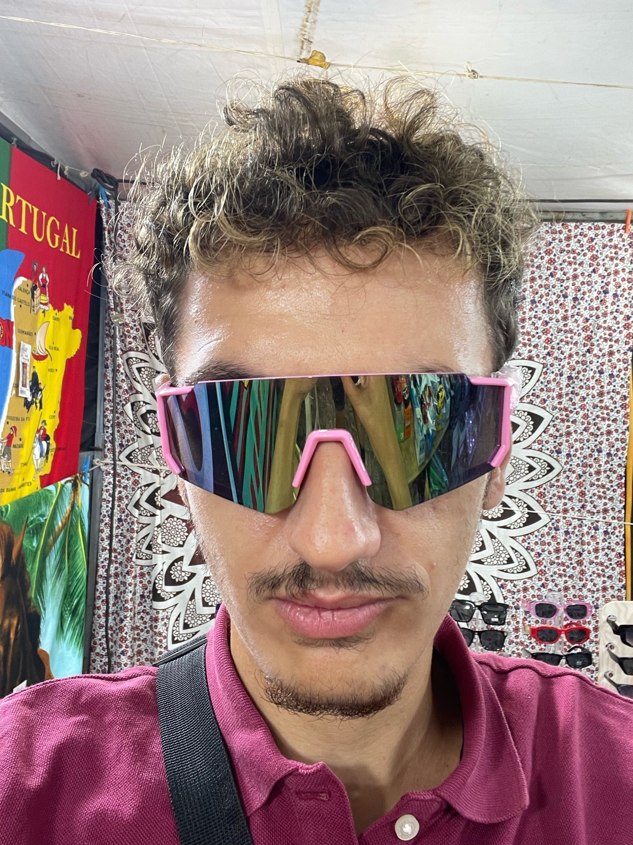 A man wearing pink sunglasses takes a selfie. The background is a shop with various items.