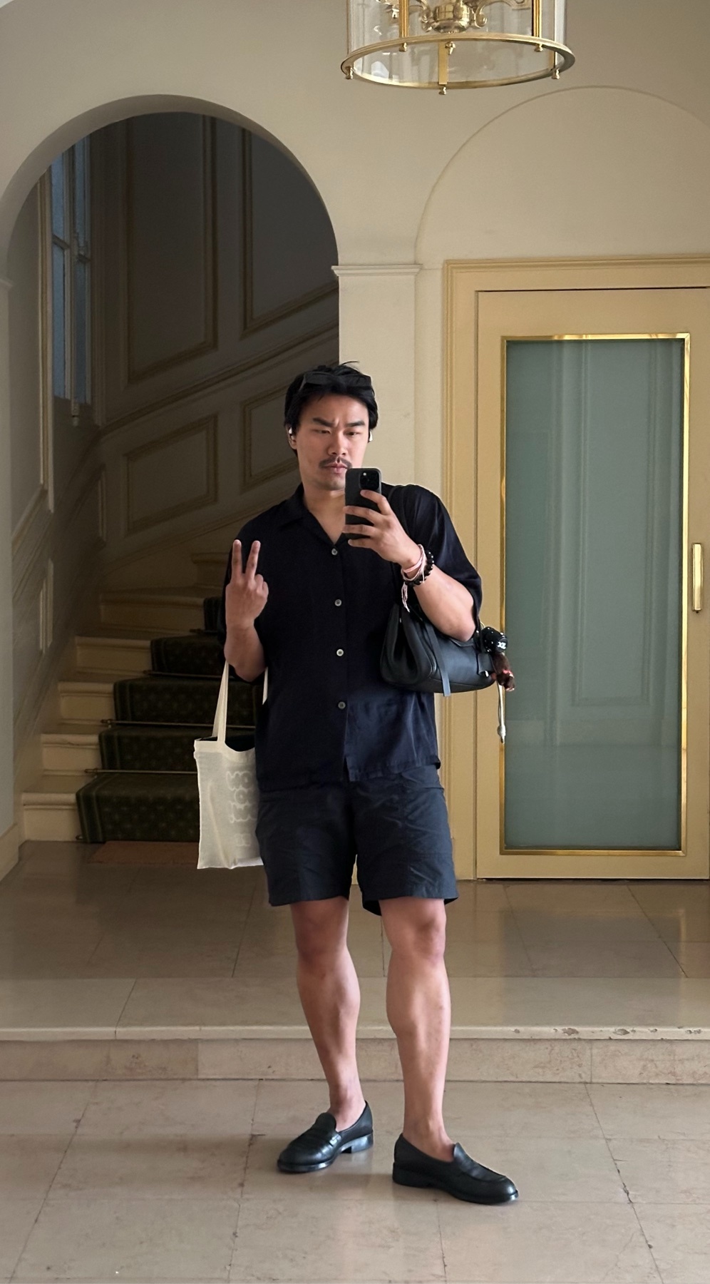 A man takes a selfie indoors, holding up two fingers in a peace sign. He is wearing a black short-sleeved button-up shirt, black shorts, and black loafers, and is carrying a tote bag and a small black purse.