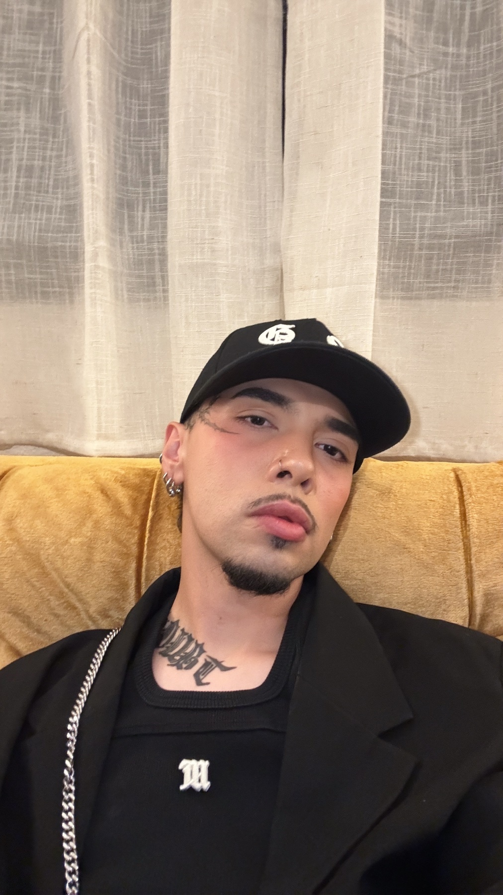 A man wearing a black baseball cap, blazer, and a black t-shirt with a white logo. He has tattoos on his neck and face, and piercings in his ears.