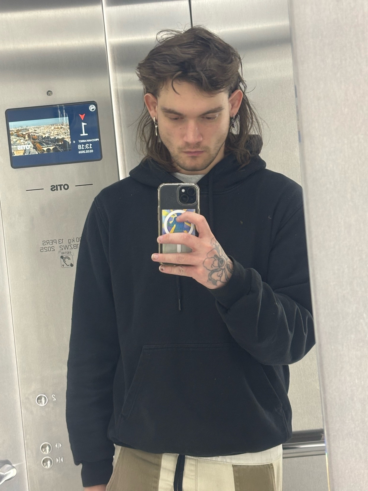 A man takes a mirror selfie in an elevator. He has a mullet and is wearing a black hoodie.