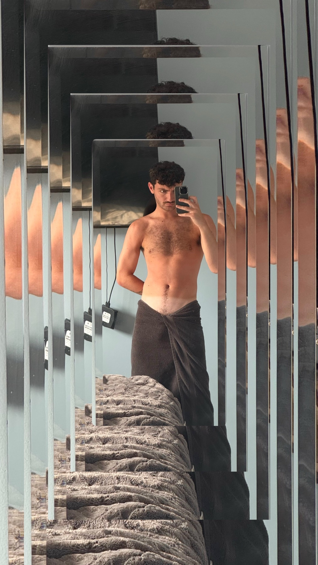 A shirtless man wearing a towel around his waist takes a mirror selfie in a bathroom. The reflection is distorted by multiple mirrored surfaces, creating a tunnel effect.