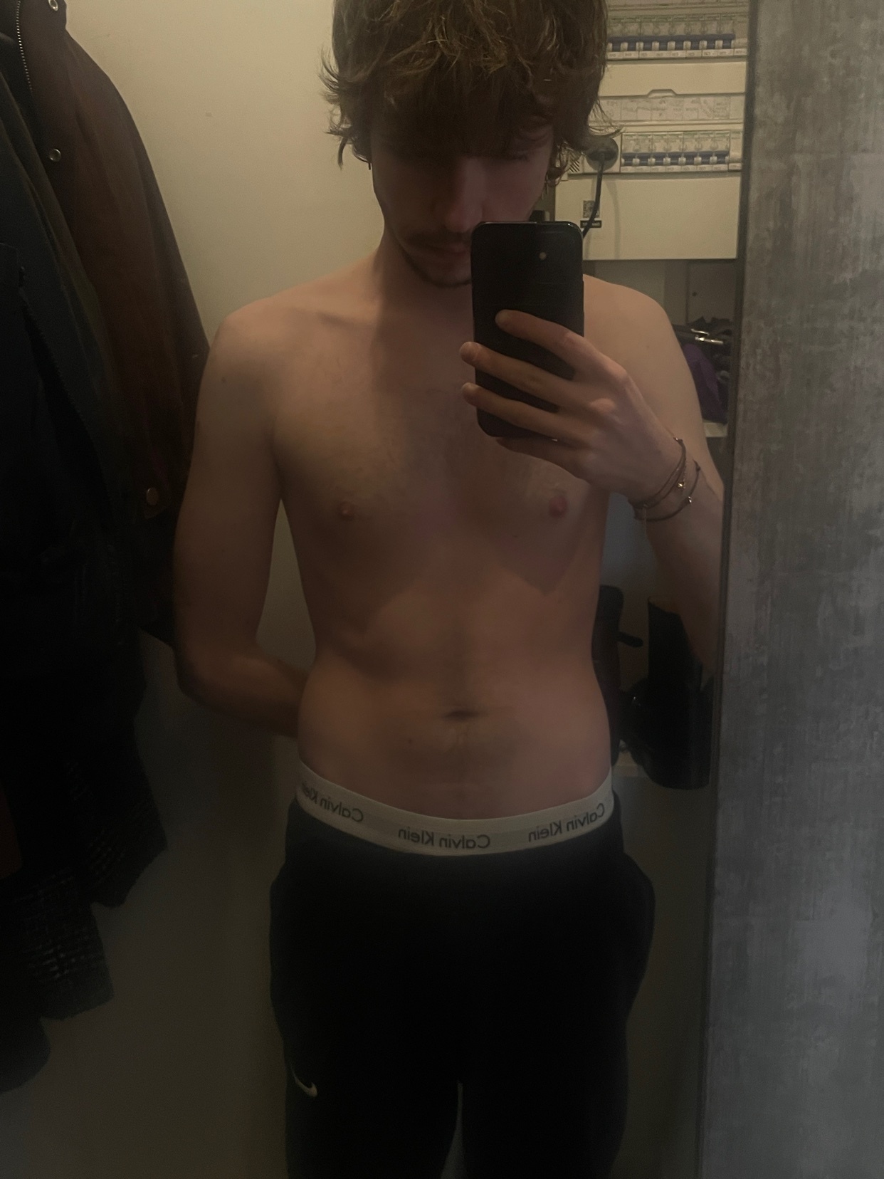 A man takes a mirror selfie, shirtless, showcasing his physique. He is wearing black pants and Calvin Klein underwear.