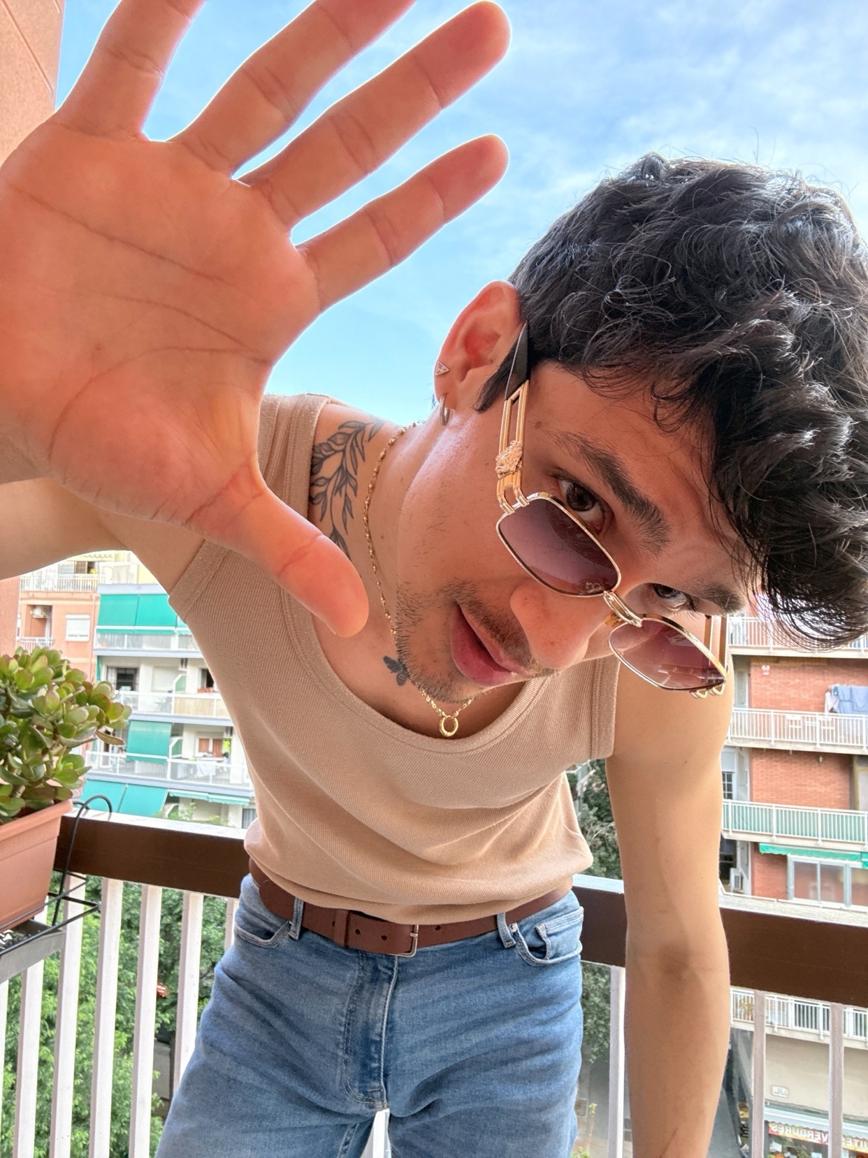 A man wearing sunglasses and a tank top poses for a selfie on a balcony, reaching his hand towards the camera.