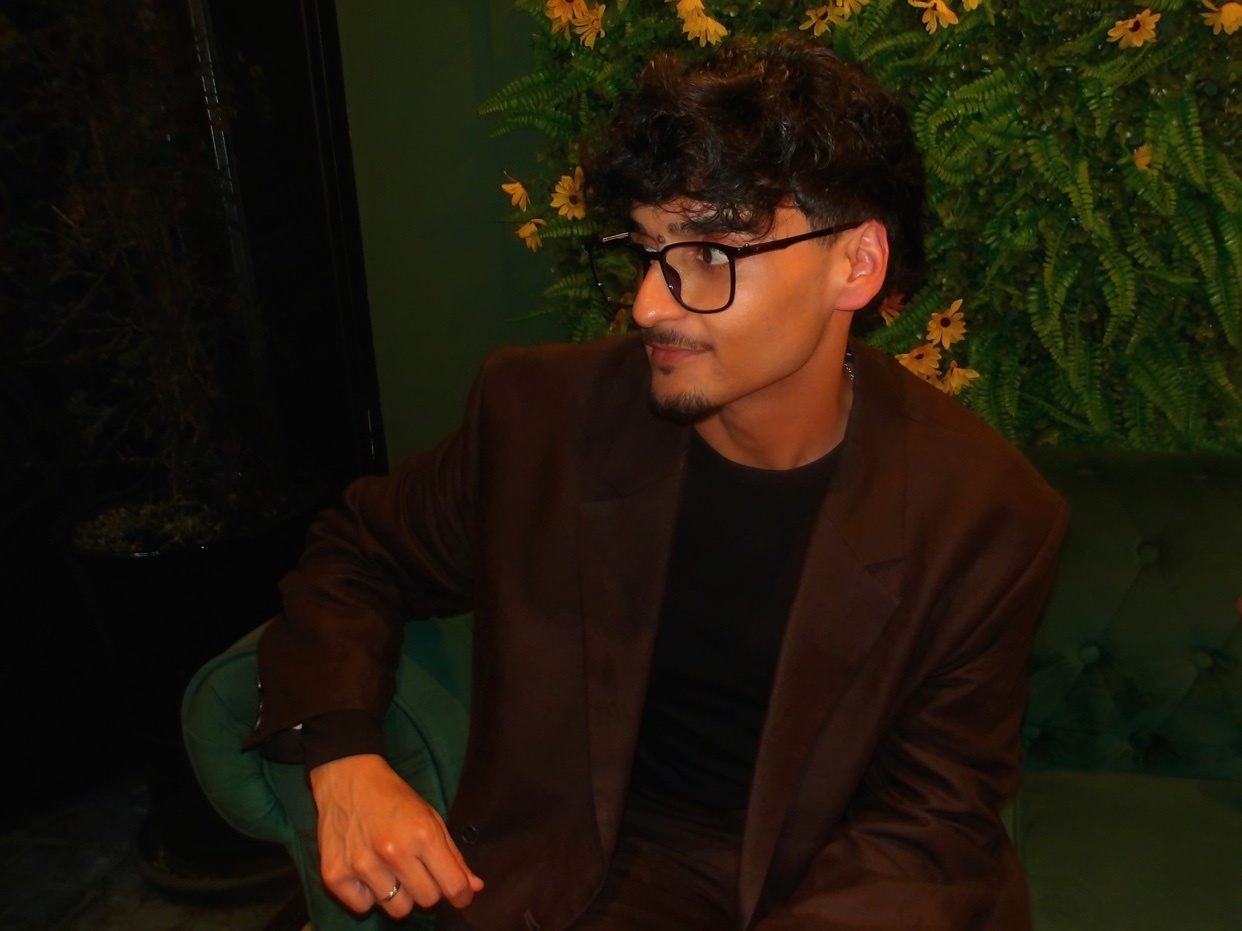 A man with dark, curly hair and glasses looks away from the camera while wearing a brown suit jacket over a black t-shirt.
