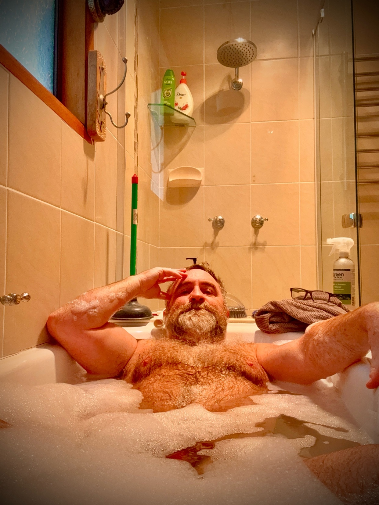 A man with a full beard and chest hair relaxes in a bubble bath, leaning back against the side of the tub with his eyes closed.