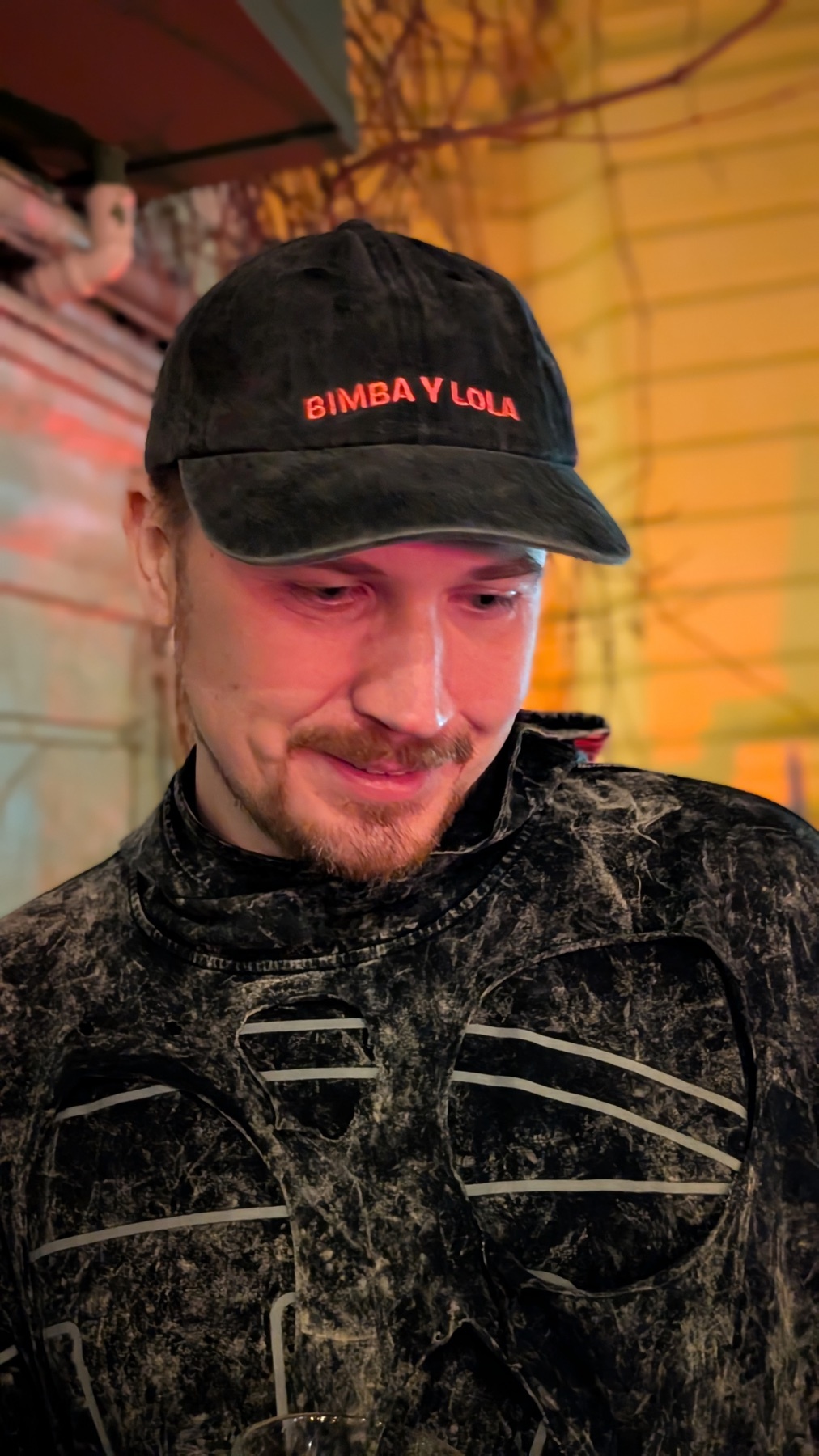 A man wearing a black cap with red text. He is wearing a patterned jacket.