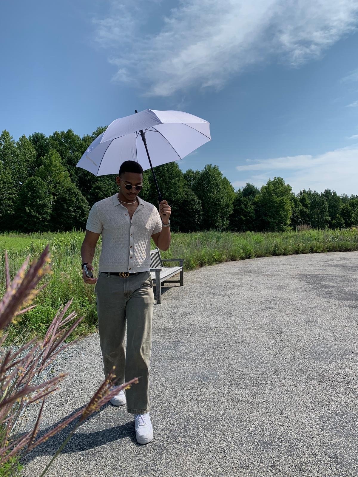A man walks down a path holding a white umbrella on a sunny day. He is wearing sunglasses and a short-sleeved collared shirt with olive green pants.