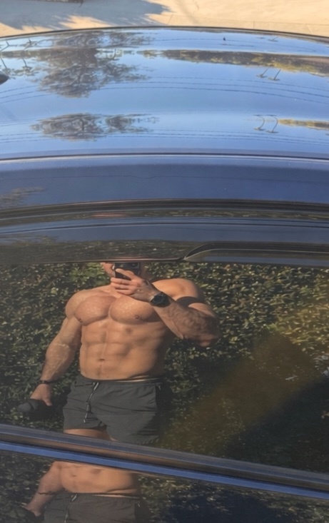 A man takes a shirtless selfie, showcasing his muscular physique. The reflection in the car window captures the man's impressive build.