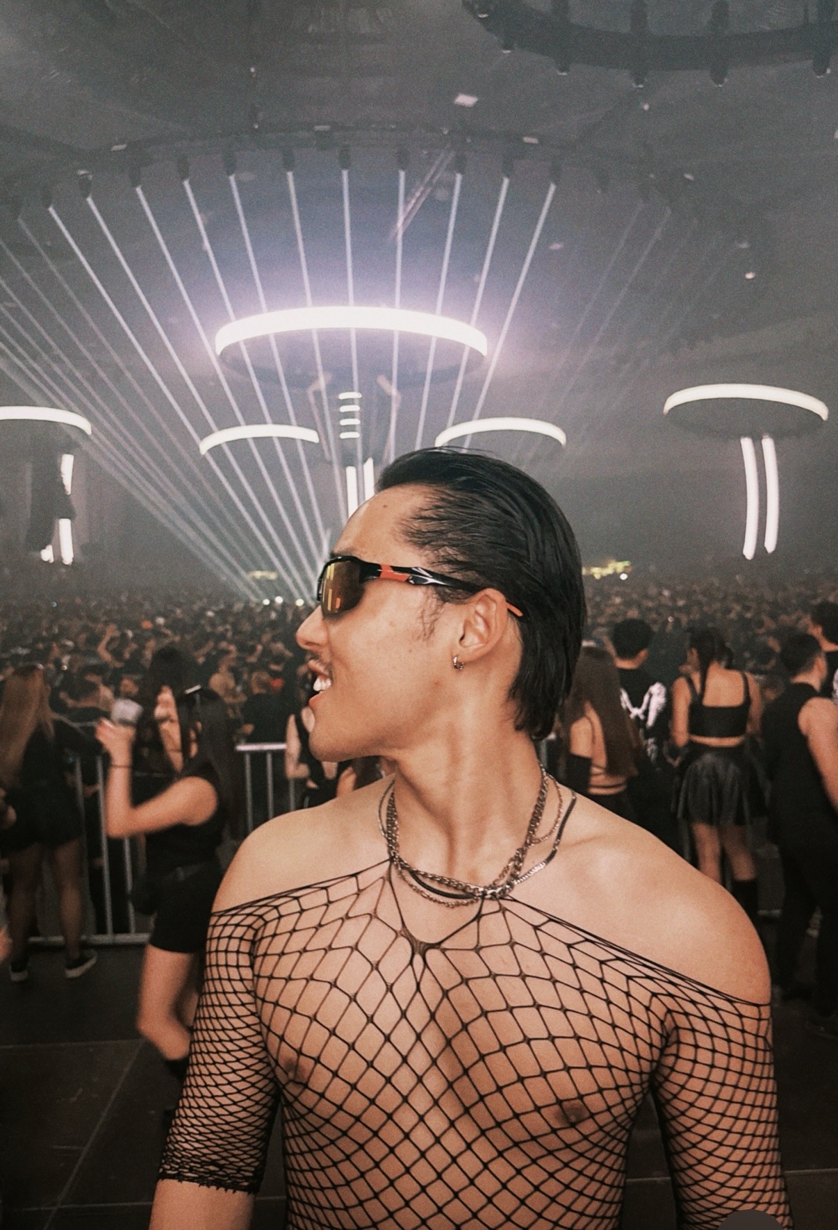 A man in a fishnet top and sunglasses poses at a concert with a large crowd in the background.