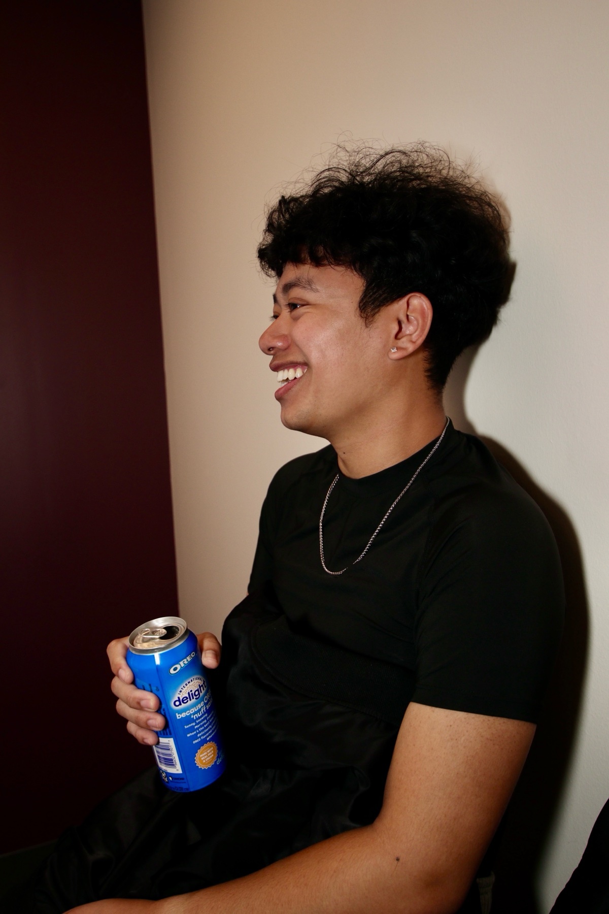 A young Asian man with curly hair smiles while holding a blue can of drink. He is wearing a black t-shirt and a silver chain.