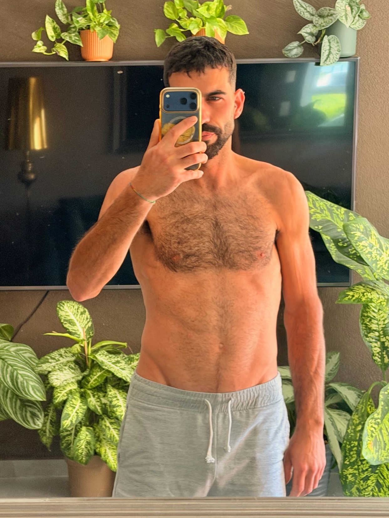 A man takes a selfie in front of a television and several potted plants. He is shirtless and wearing grey sweatpants.