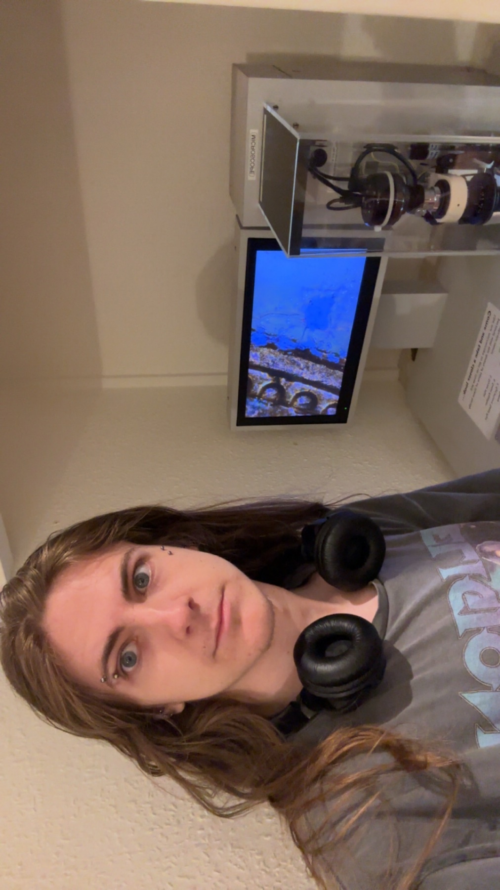 A person with long brown hair and headphones around their neck takes a selfie in front of a display featuring a microscope.