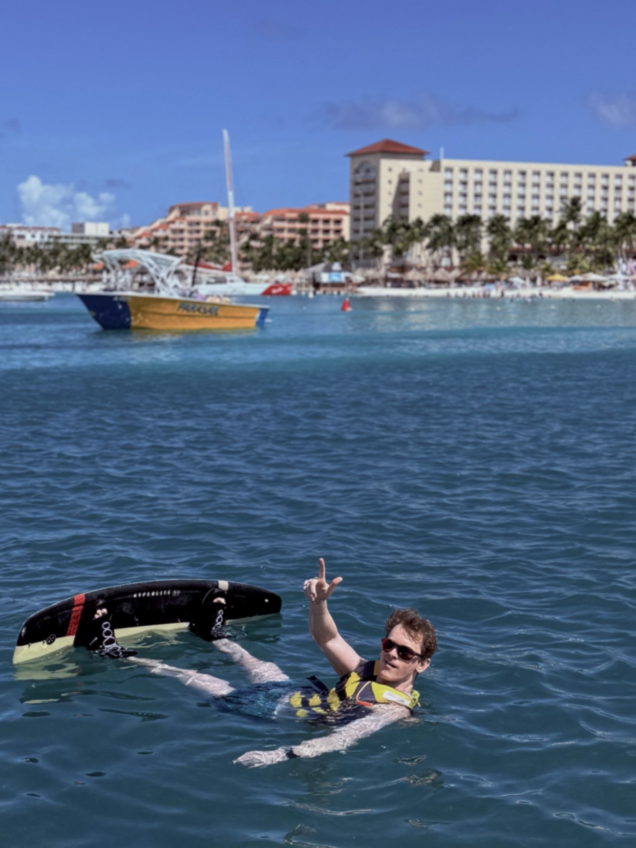 A man in sunglasses and a life vest floats in the ocean with a wakeboard. A boat and buildings are visible in the background.