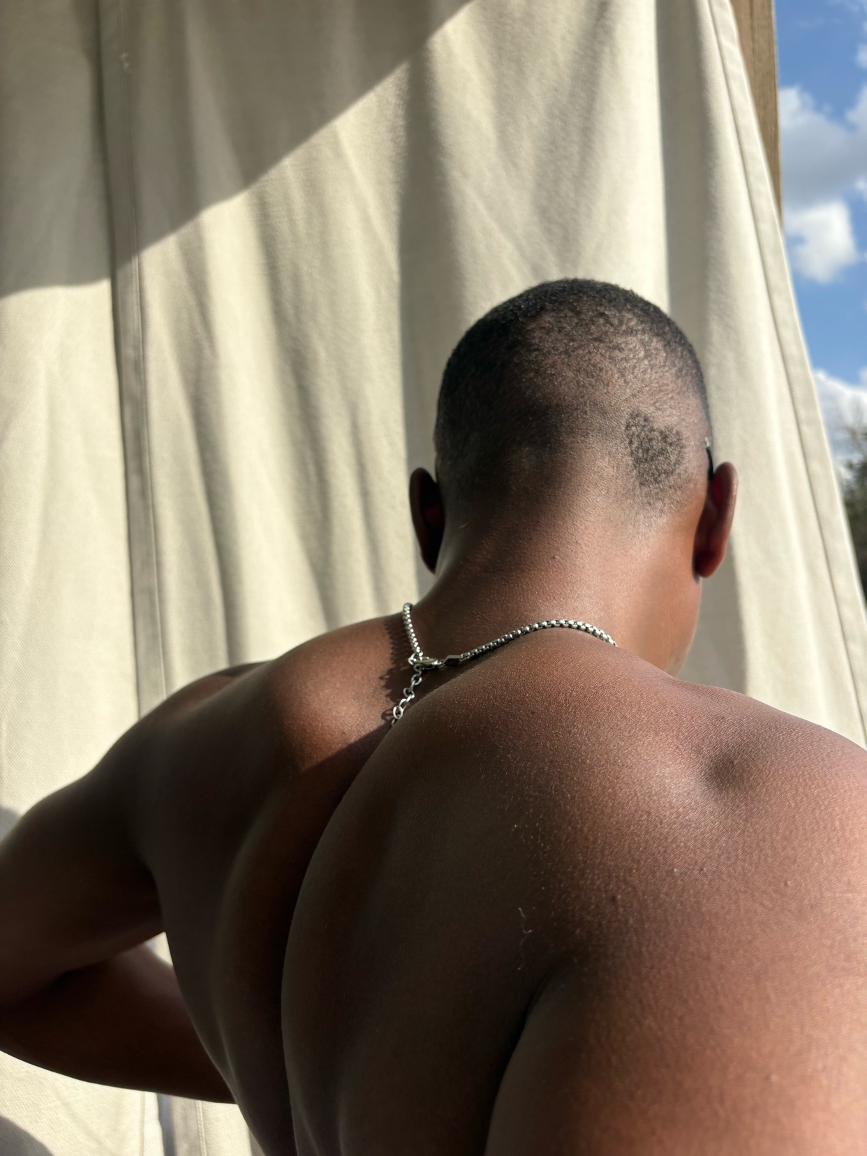A close-up shot of a man's back, showing off his muscular physique and a small tattoo on the back of his head. He is wearing a silver chain necklace.