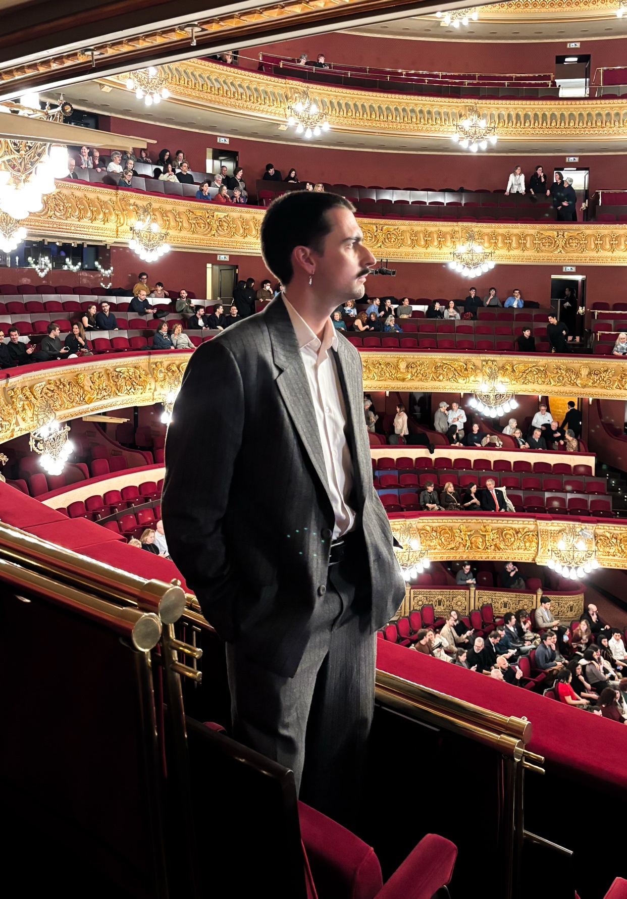 A man in a suit stands in a theater, looking out at the audience. The theater's architecture is ornate and visually interesting.
