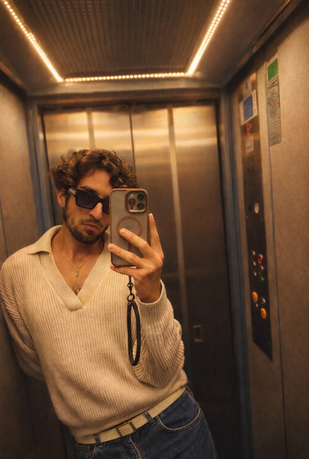 A man with curly hair and sunglasses takes a selfie in an elevator, wearing a collared sweater and jeans.