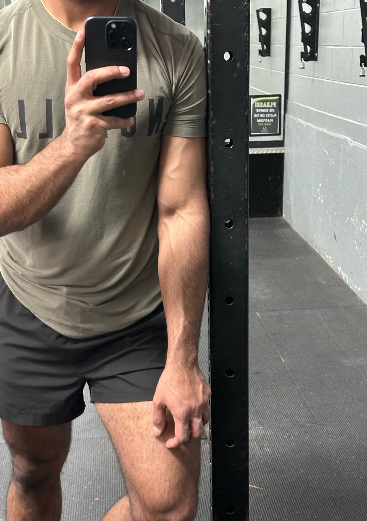 A man flexes his bicep in a gym setting. He is shirtless and wearing shorts.
