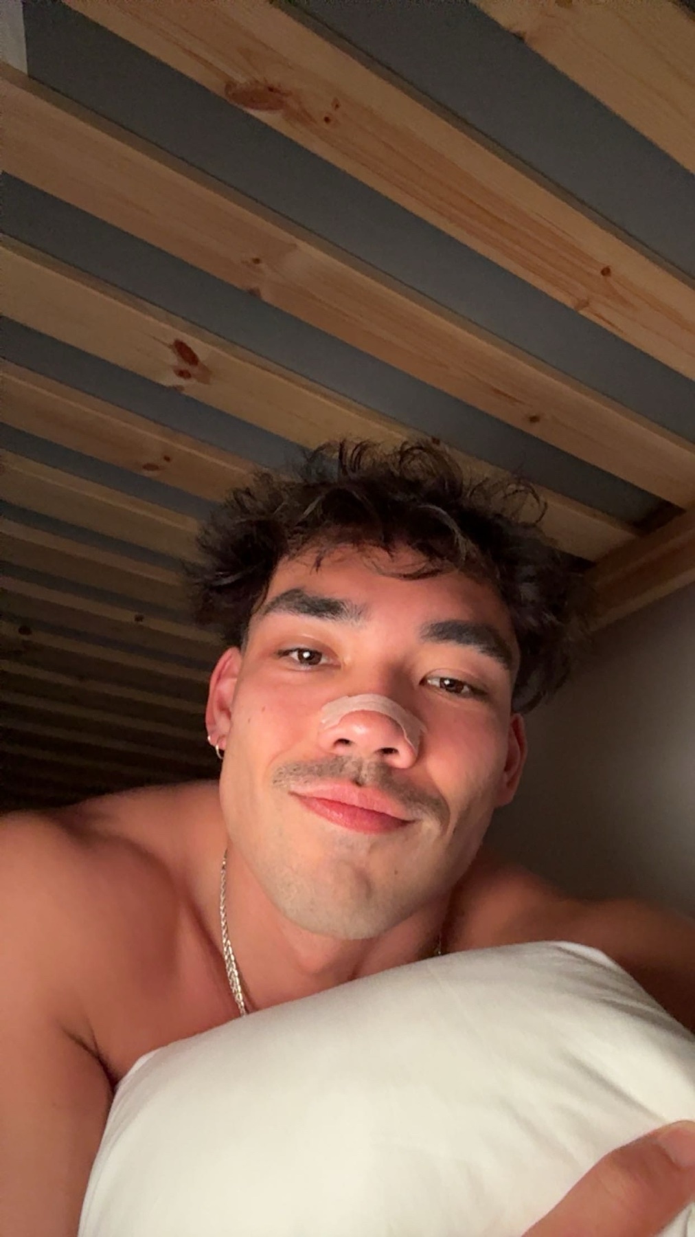 A close-up selfie of a man lying in bed, smiling at the camera. He has dark, curly hair, stubble, and is wearing a necklace.