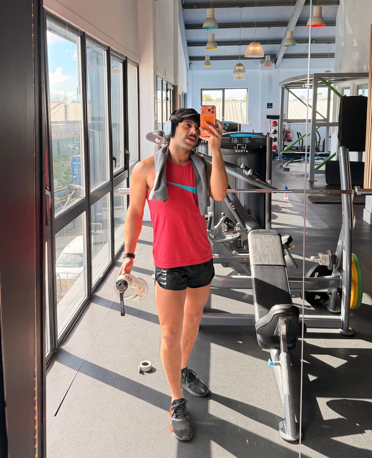 A man takes a selfie in a gym, holding a water bottle and wearing a red tank top, black shorts, and a towel draped over his shoulders. He appears to be finished with a workout, with gym equipment visible in the background.