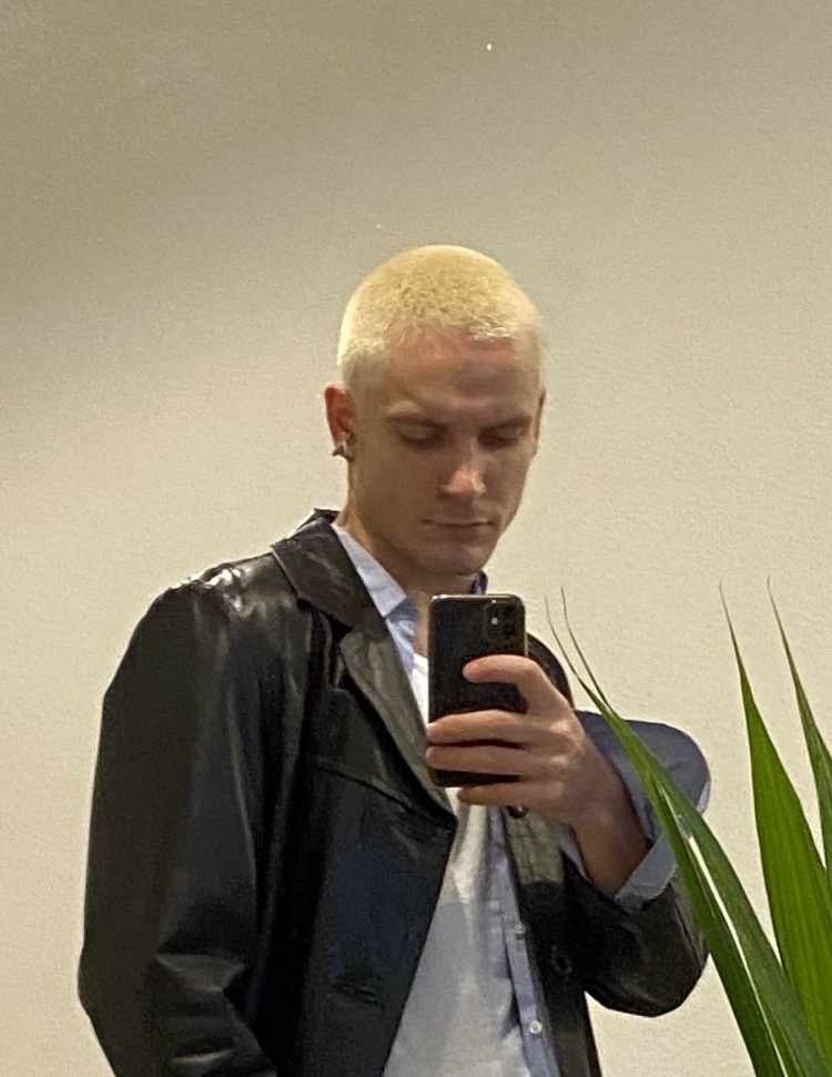 A man with short, blond hair takes a selfie in a mirror, wearing a black leather jacket over a light blue button-up shirt. A plant is visible in the foreground.