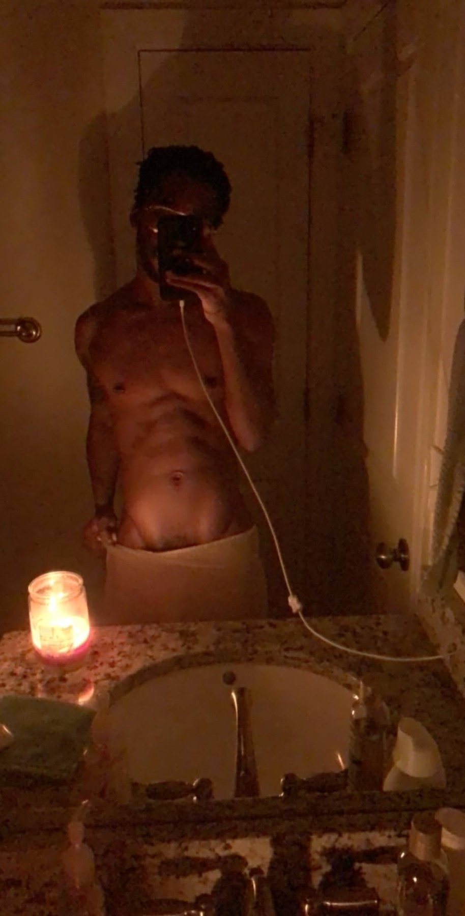 A man takes a mirror selfie in a dimly lit bathroom, wearing a towel around his waist. A lit candle sits on the counter next to the sink.