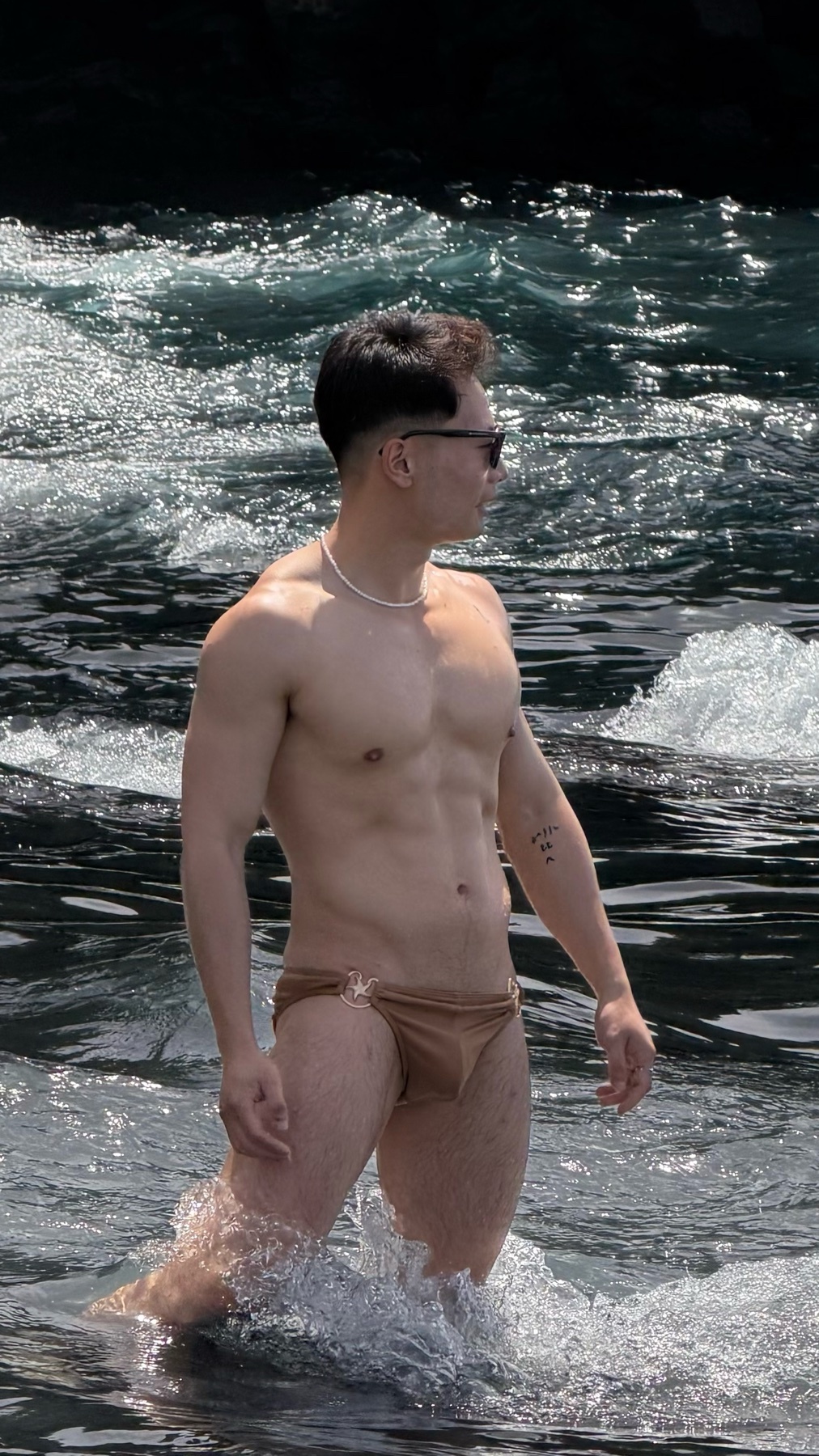 A shirtless man with sunglasses stands in the water wearing swimwear. He has a muscular physique and appears to be of Asian descent.
