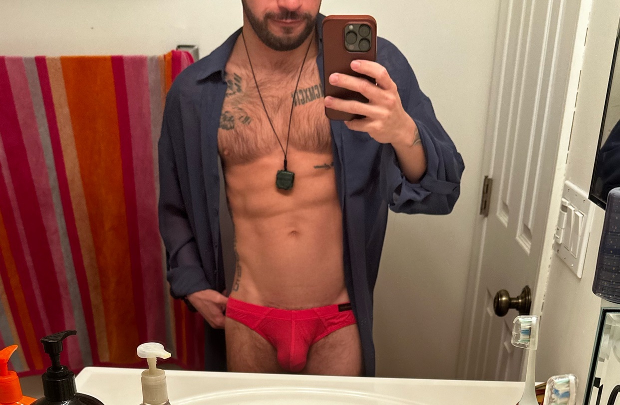 A man takes a mirror selfie, shirtless and wearing red underwear. He has a muscular physique and visible tattoos.