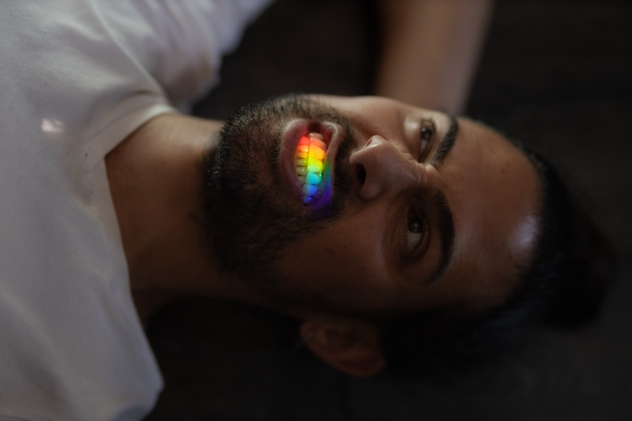 A close-up shot of a man with a beard and dark hair, lying down. His mouth is open, revealing teeth colored like a rainbow.