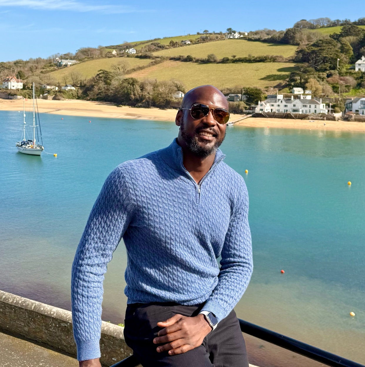 A man poses for a photo outdoors. He is wearing sunglasses and a blue sweater, with a scenic landscape in the background.