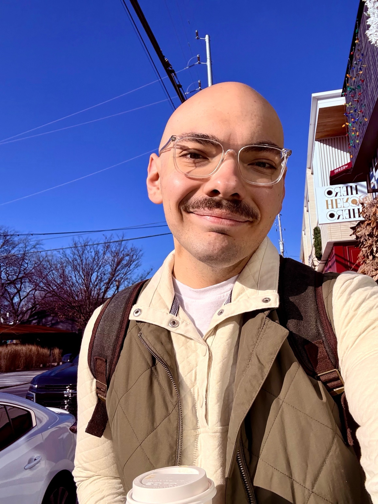 A man with a bald head and mustache smiles at the camera while holding a coffee cup. He is wearing glasses and a stylish outfit.