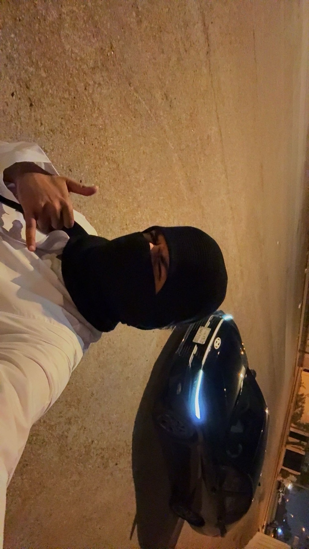 A person wearing a black balaclava and a white thobe poses for a selfie next to a scooter at night.