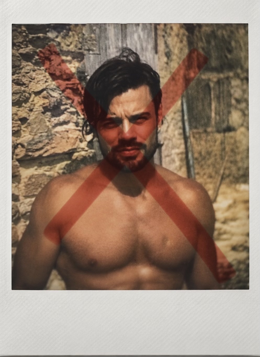 A shirtless man with a beard and dark hair is pictured in a polaroid photo. A large red X is overlaid on the image.