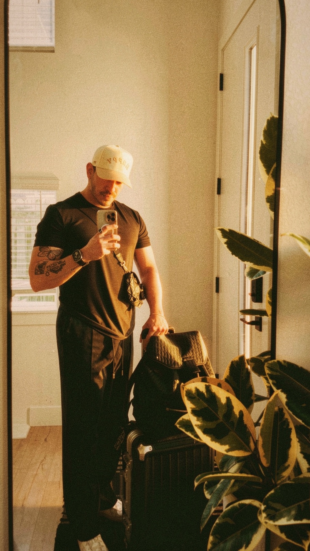 A man in a black t-shirt and hat takes a selfie in a mirror, holding a phone and a backpack. He is standing next to a suitcase, with a large plant to his right.