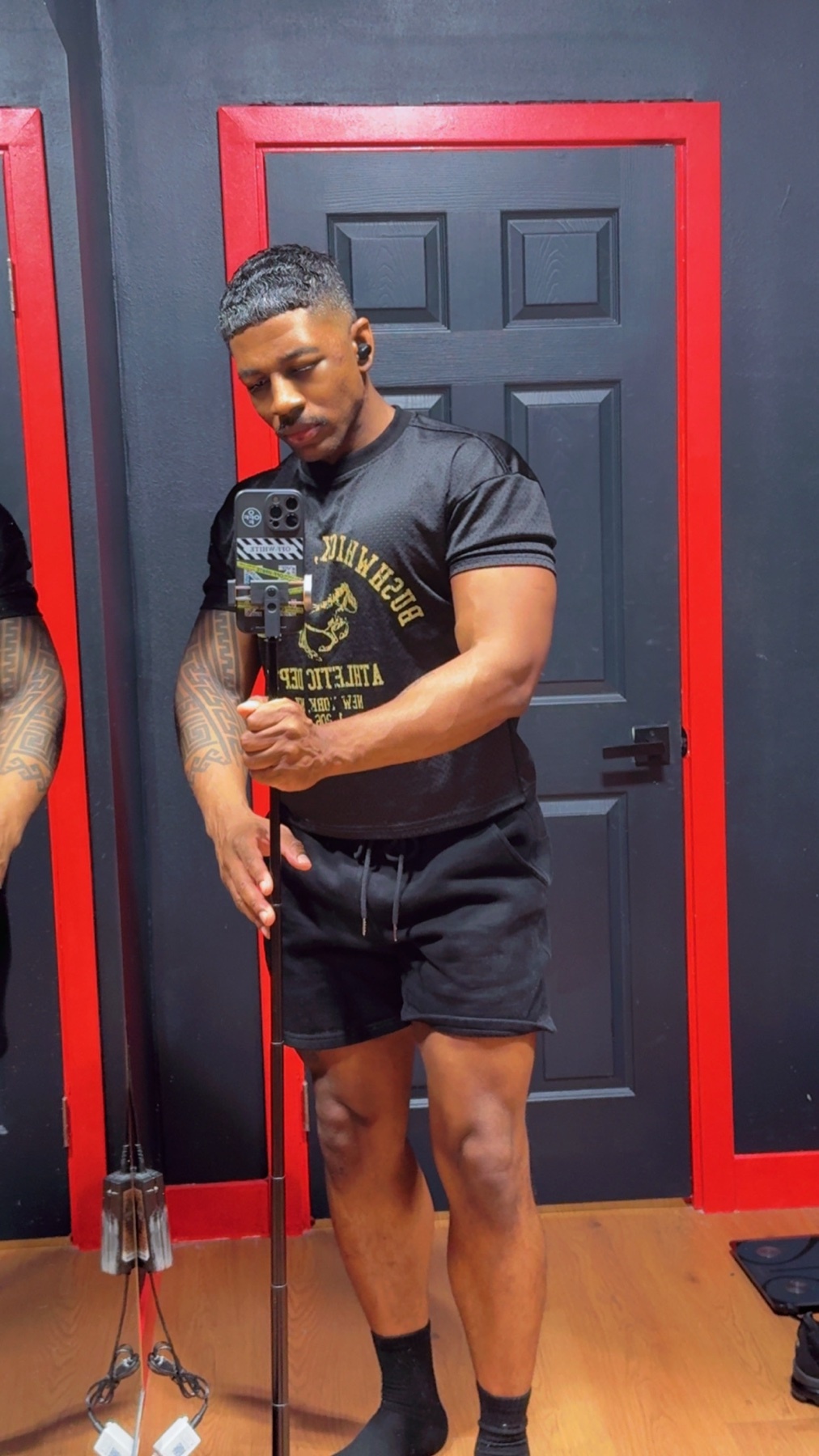 A man with tattoos on his arms poses for a mirror selfie in a gym. He is wearing a black t-shirt and black shorts.