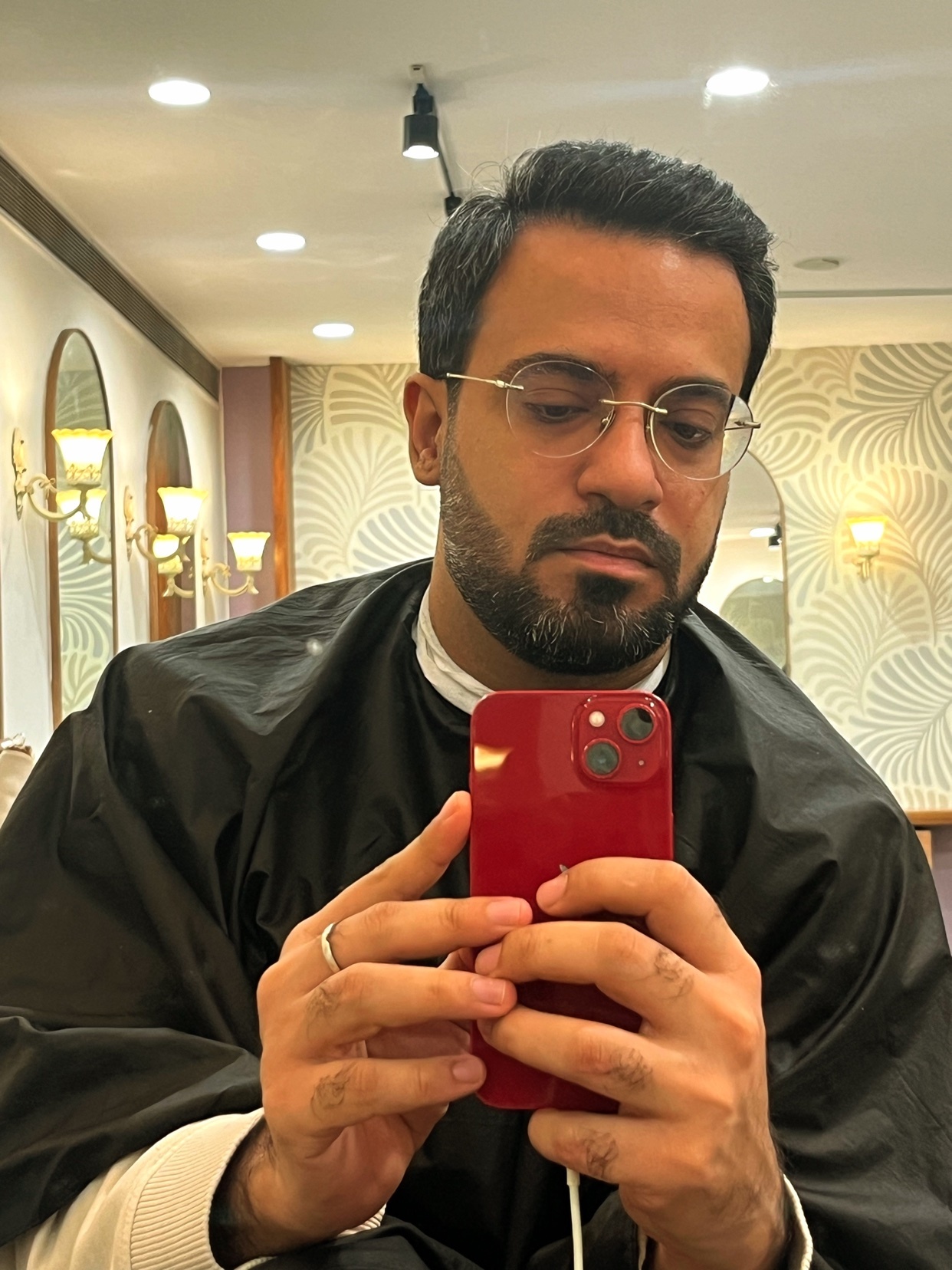 A man takes a selfie at a salon. He is wearing glasses and a black cape.