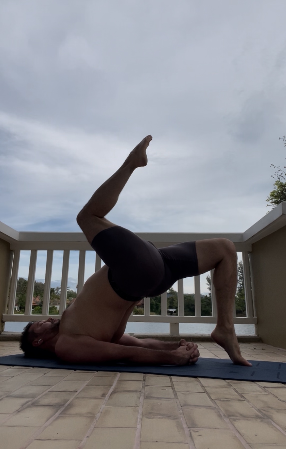 A man performs a yoga bridge pose outdoors. He is shirtless and wearing shorts.