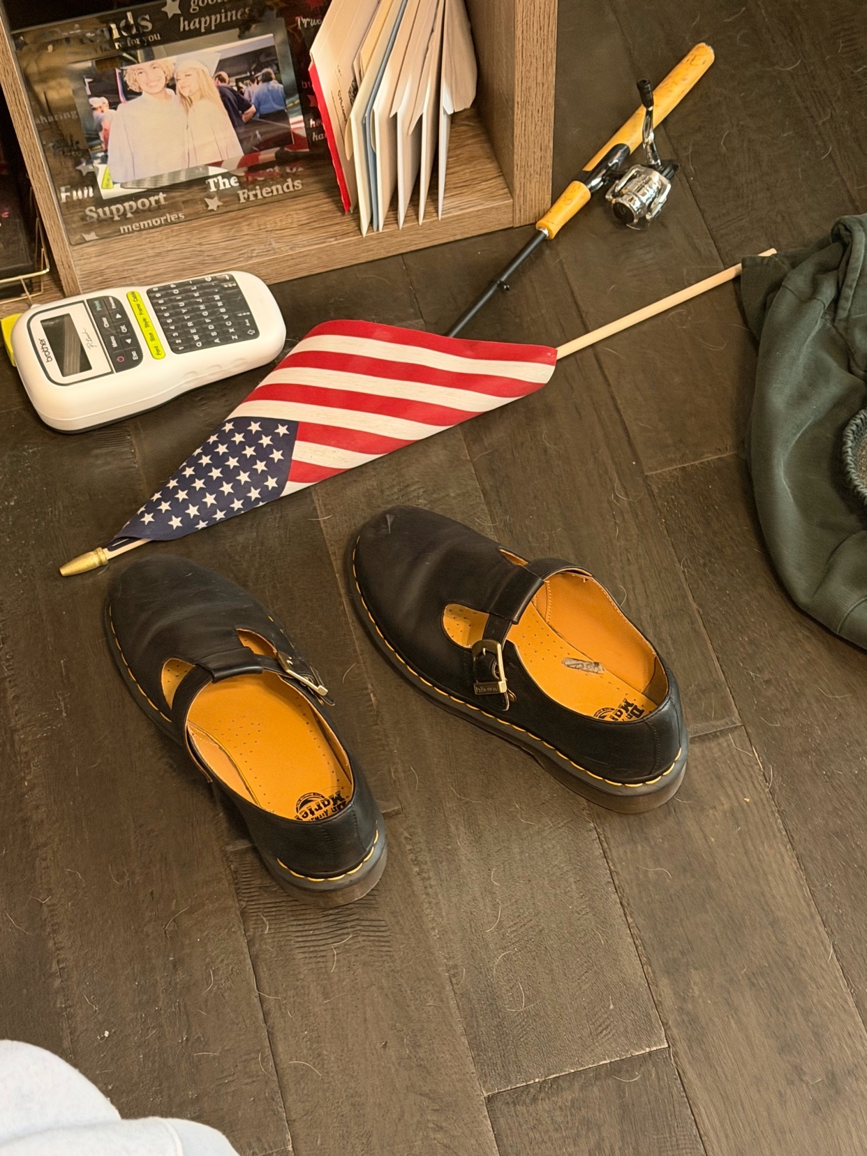 A pair of black Mary Jane shoes are placed on a dark wood floor next to an American flag, a fishing rod, a label maker, and some books.